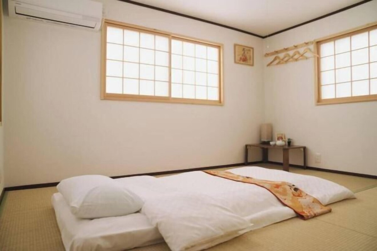 Ein Zimmer mit Tatami-Boden, einem Bett mit weißer Bettwäsche, zwei Fenstern mit Holzrahmen und einem kleinen Tisch mit einer Lampe.
