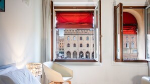 Una habitació d'hotel amb un llit, una cadira, una finestra amb cortina vermella i vista a un edifici històric.