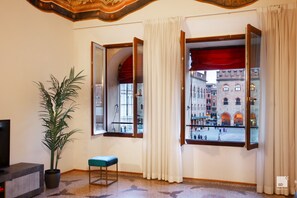 Une pièce dotée d’une grande fenêtre donnant sur un bâtiment historique et d’un balcon. On y trouve une plante en pot et un banc près de la fenêtre.