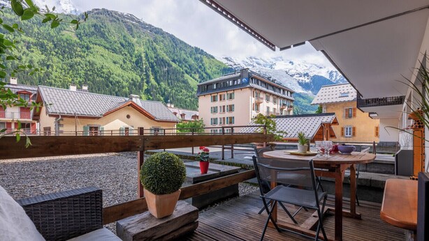 A wooden deck with a table and chairs, overlooking a mountainous landscape with buildings.