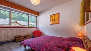 A bedroom with a large bed, a red chair, a wooden bench, a window with a view of houses, and a painting on the wall.
