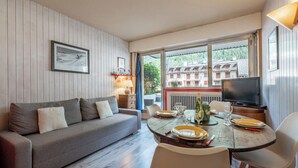 A cozy room with a sofa, dining table, and chairs. A television is on the table, and there is a window with a view of buildings outside.