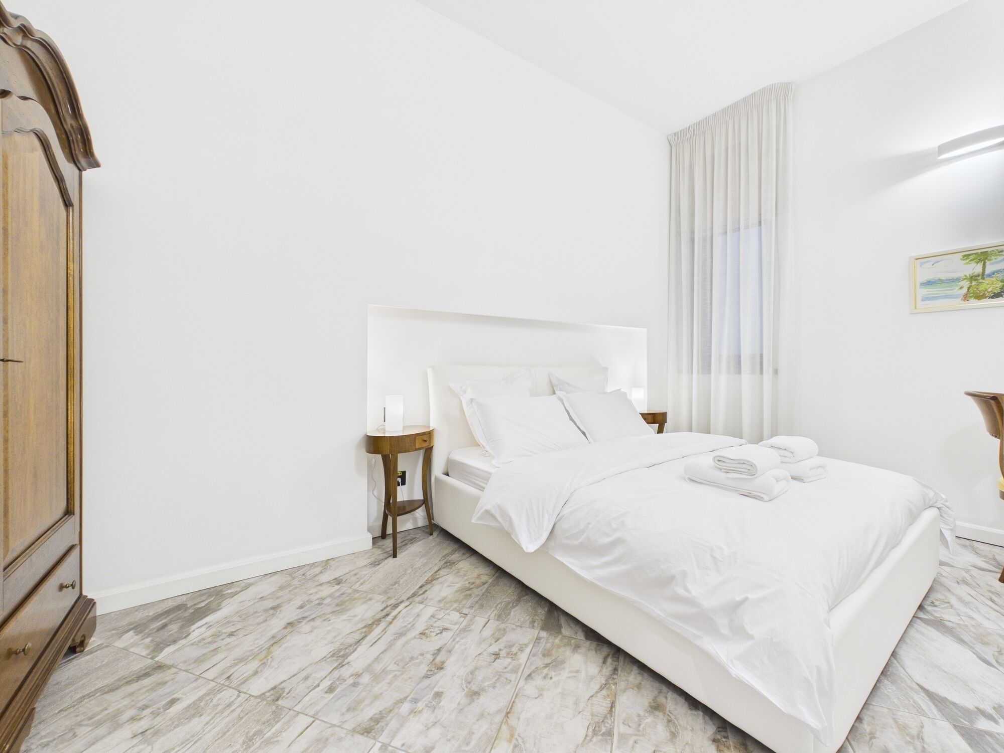 A bedroom with a bed, a nightstand, a wooden wardrobe, and a painting on the wall.