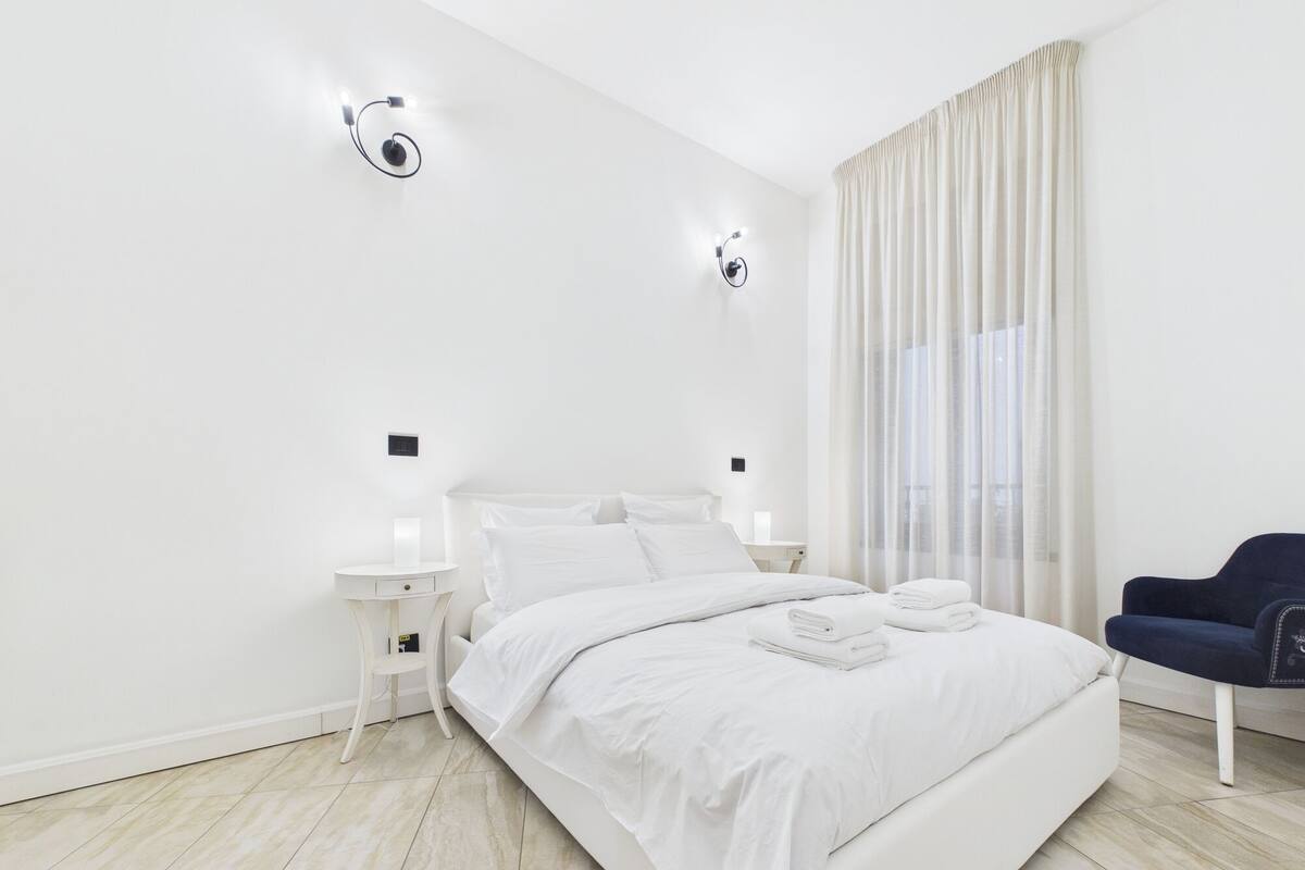 Ein modernes Hotelzimmer mit einem großen Bett, zwei Nachttischen mit Lampen, einem Sessel und einem Fenster mit Vorhängen.