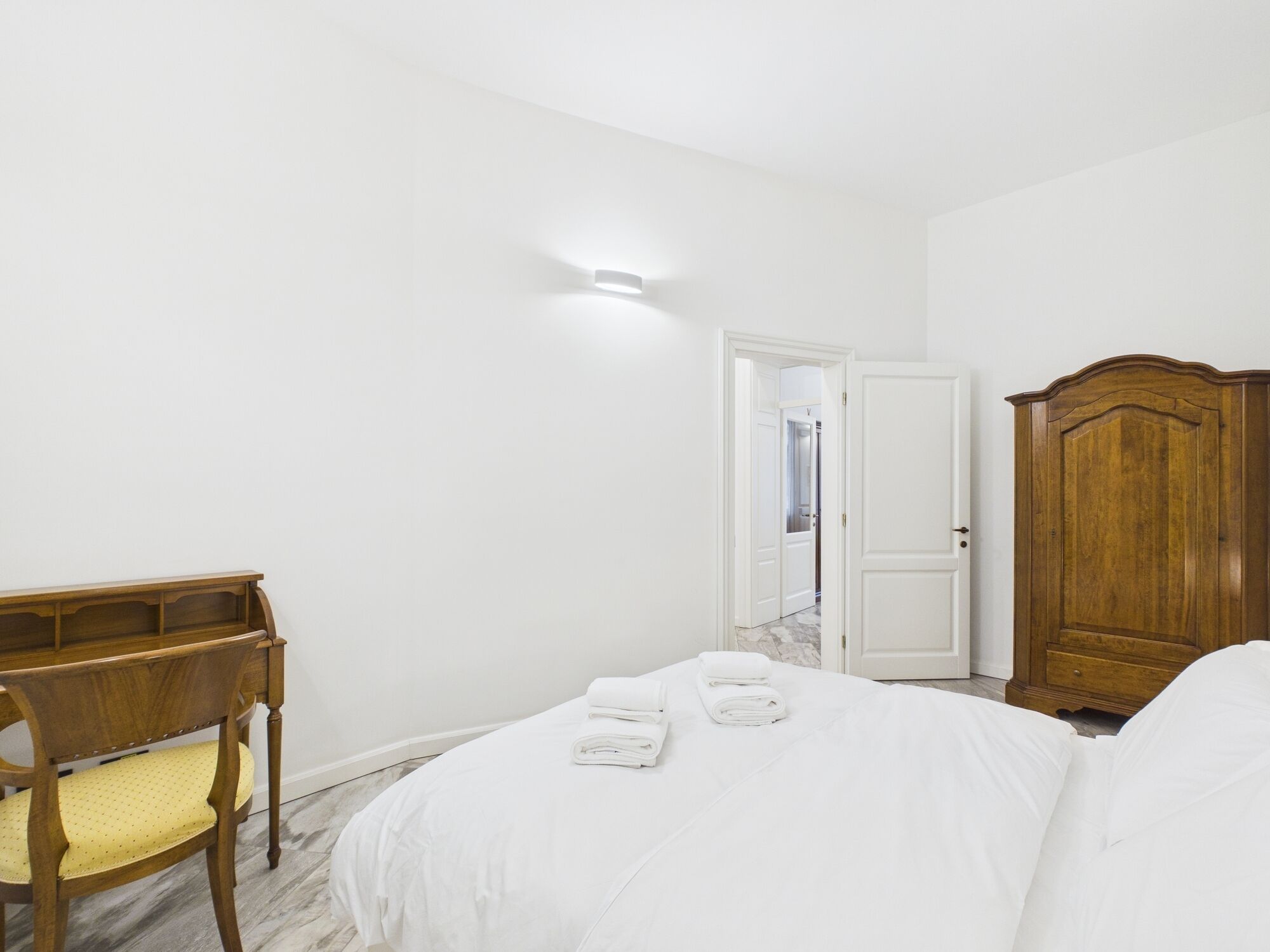 A bedroom with a bed, a wooden desk, a chair, a wardrobe, and a door leading to another room.