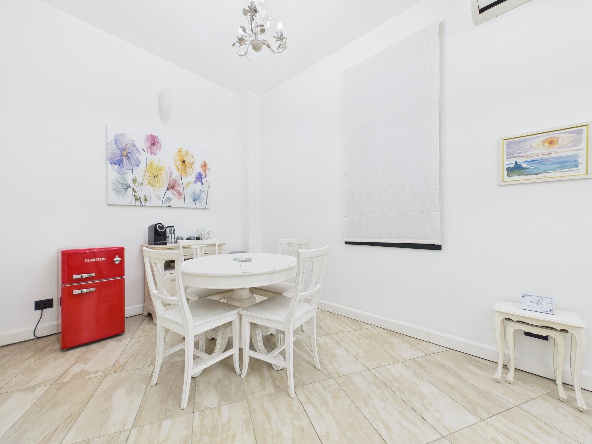 Une pièce avec une table et des chaises blanches, un meuble de rangement rouge, un tableau représentant des fleurs et une petite table sur laquelle est posé un panneau.