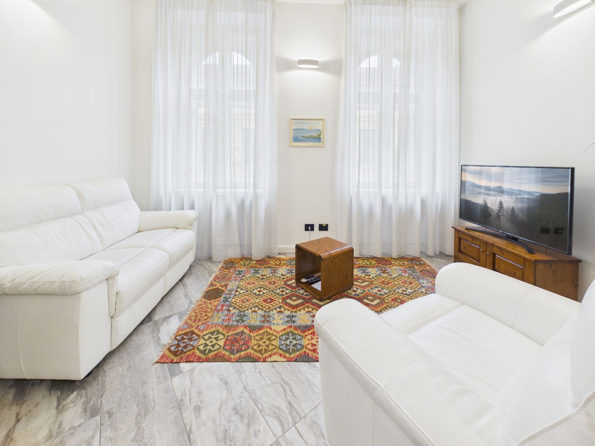 Una sala de estar con sofá blanco, televisor sobre un mueble de madera, alfombra estampada y cortinas de gasa.