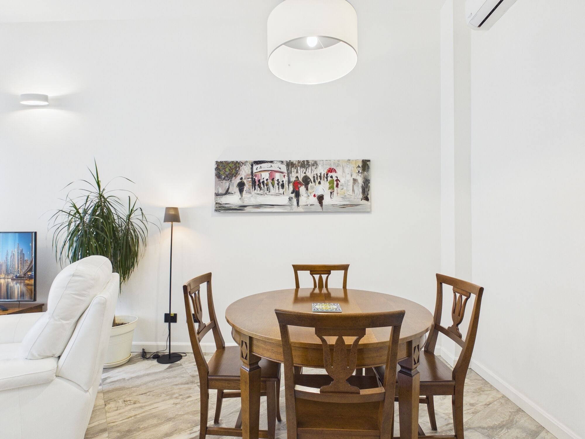 Ein Essbereich mit einem runden Holztisch und vier Stühlen, einem weißen Sofa, einer Stehlampe und einem Gemälde an der Wand.