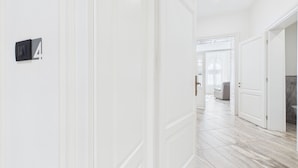 Een gang met witte muren en een marmeren vloer, die leidt naar een kamer met een grijze bank en een witte deur.