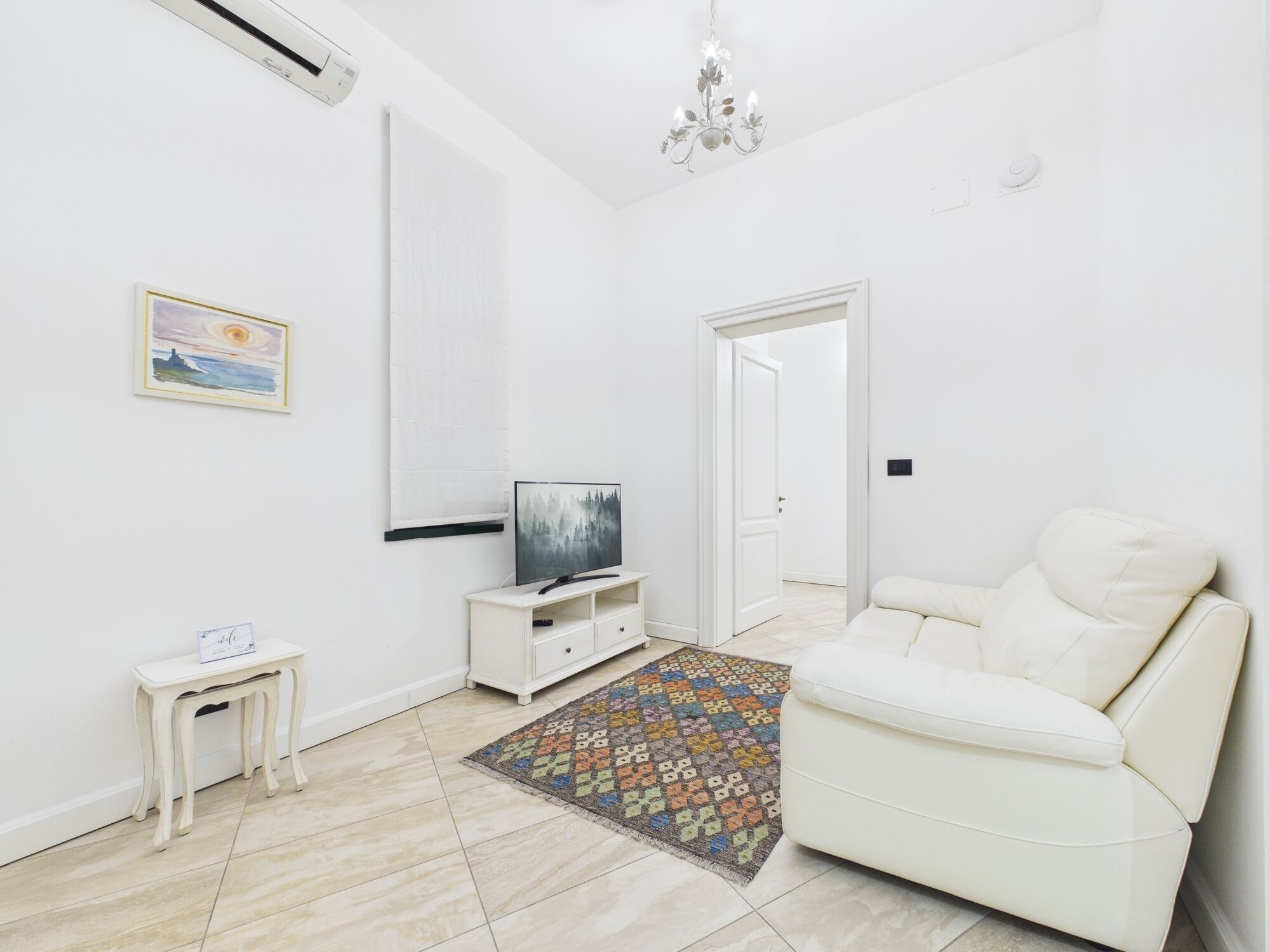 A modern living room with a white sofa, a TV on a white stand, a small table, a colorful rug, and a large mirror on the wall.