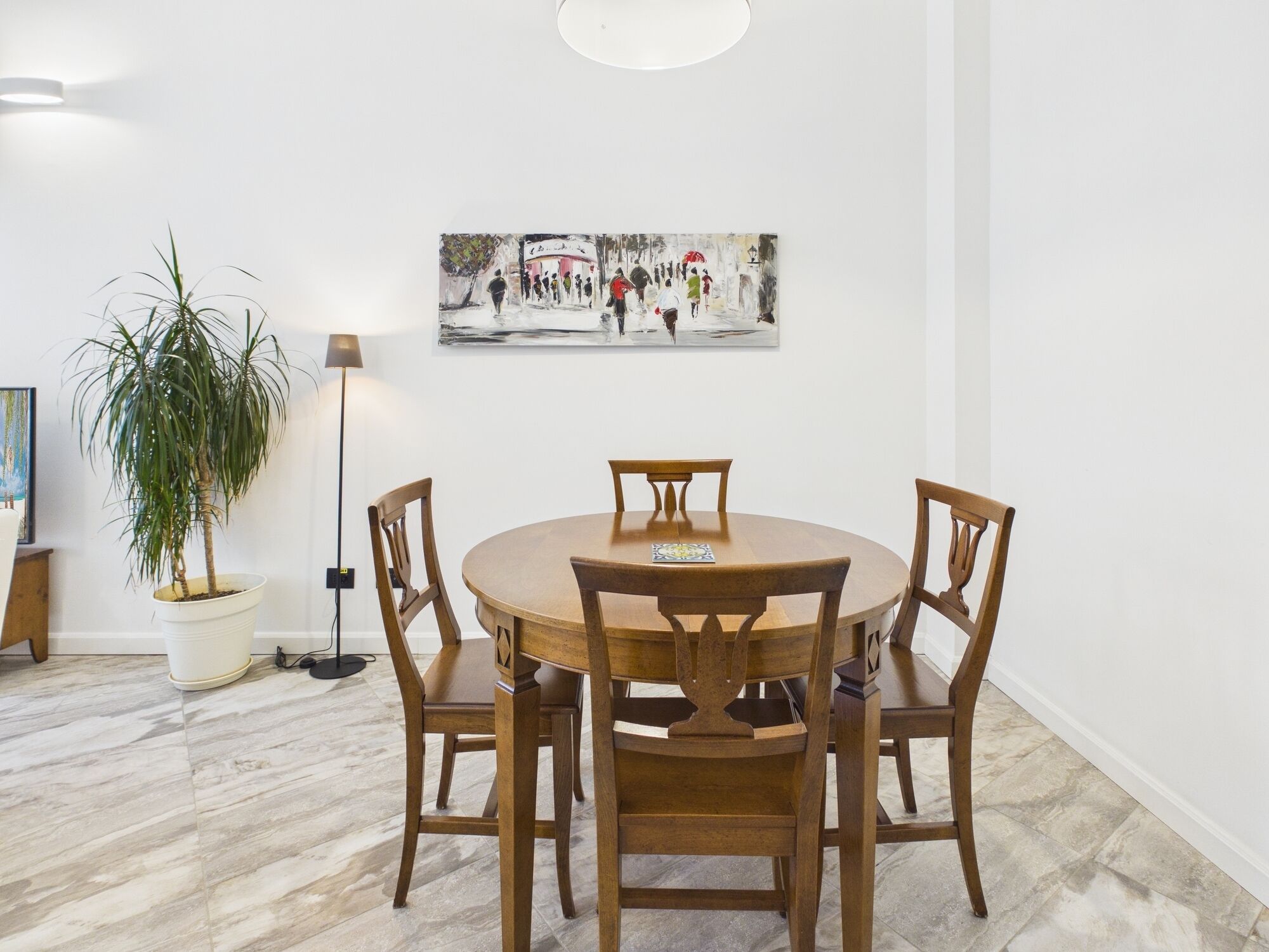 A dining area with a round wooden table and four chairs, a potted plant, and a painting on the wall.