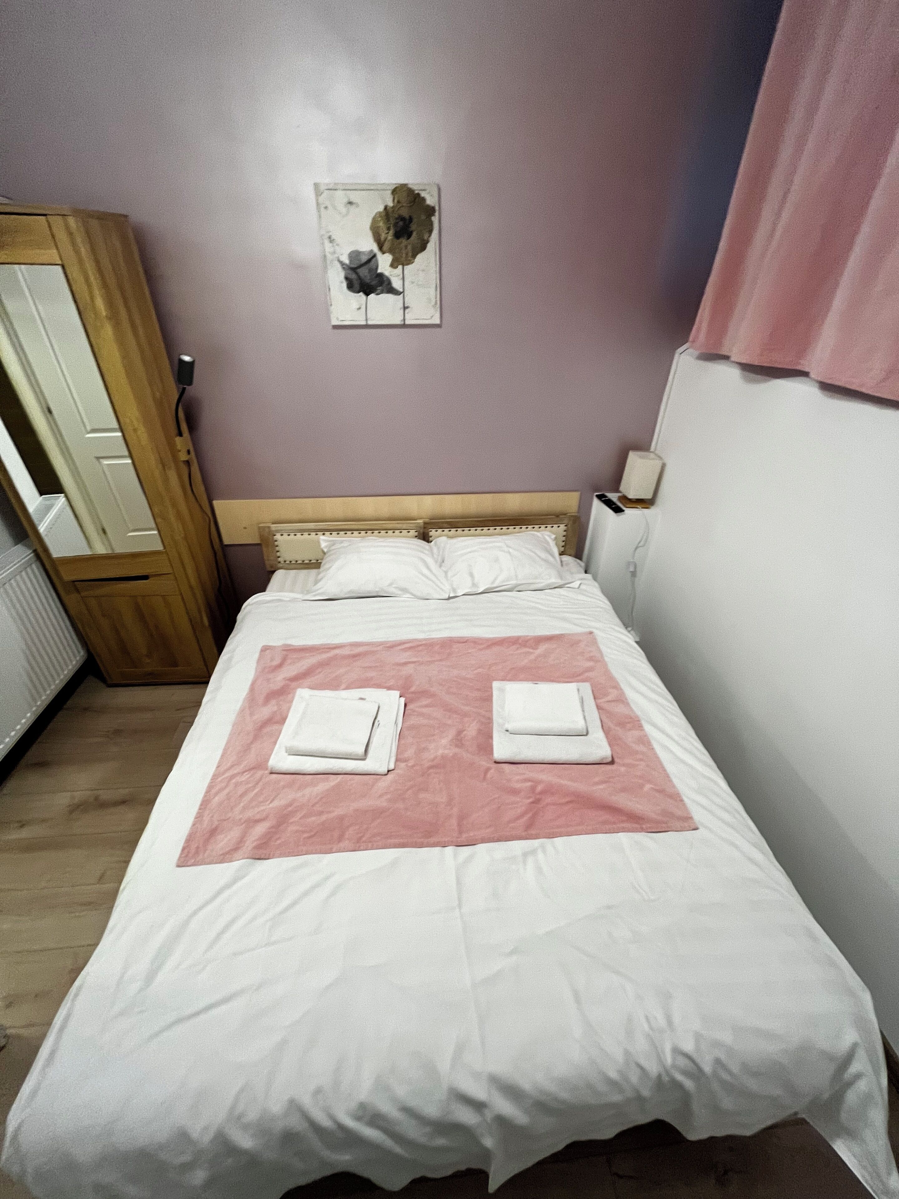 A neatly made bed with two folded white towels, a wooden headboard, a wall clock, and a flower painting.