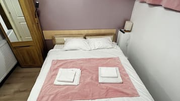 A neatly made bed with two folded white towels, a wooden headboard, a wall clock, and a flower painting.