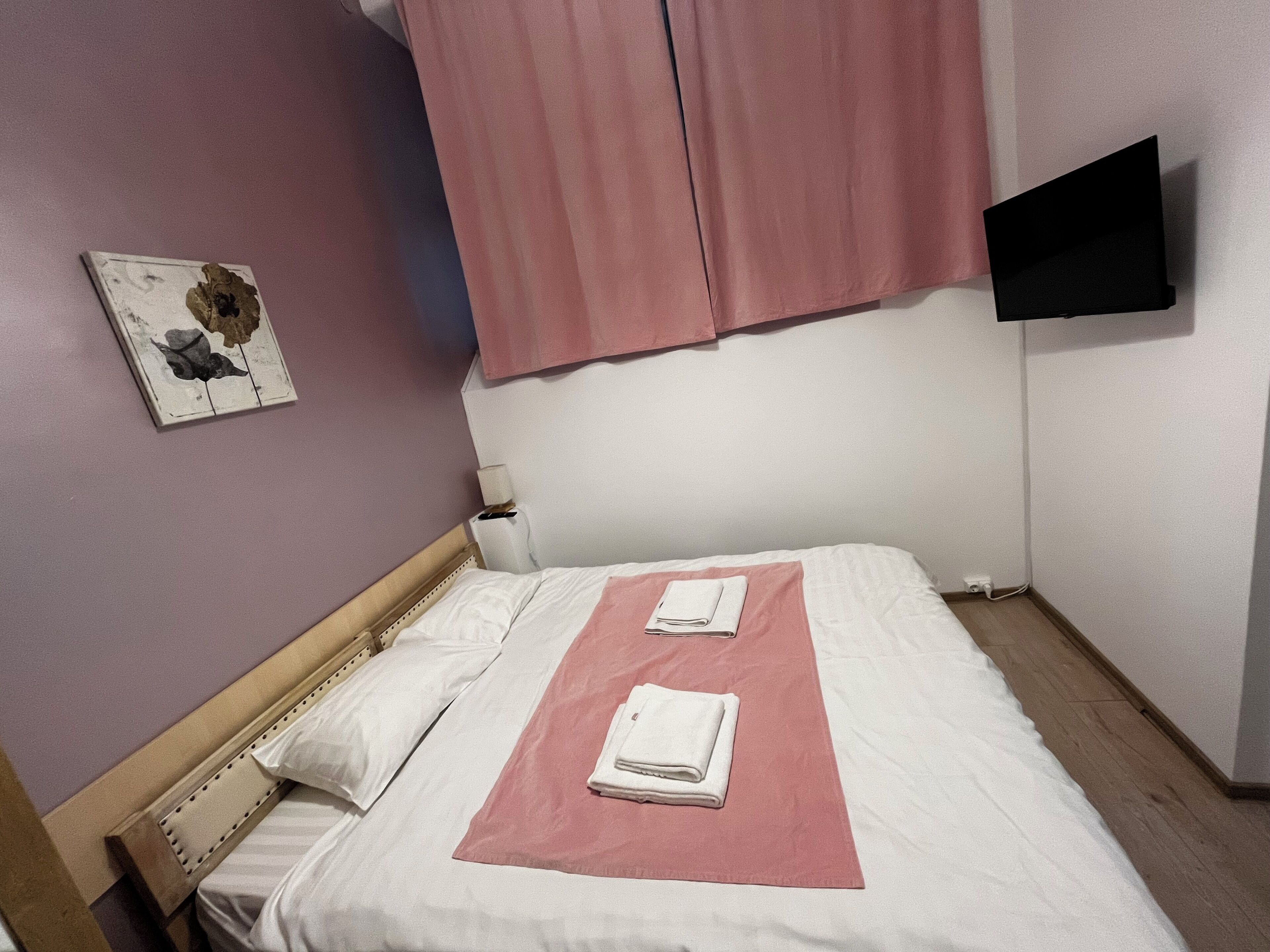 A neatly made bed with white and pink linens, a wooden headboard, a wall-mounted TV, and a small painting on the wall.