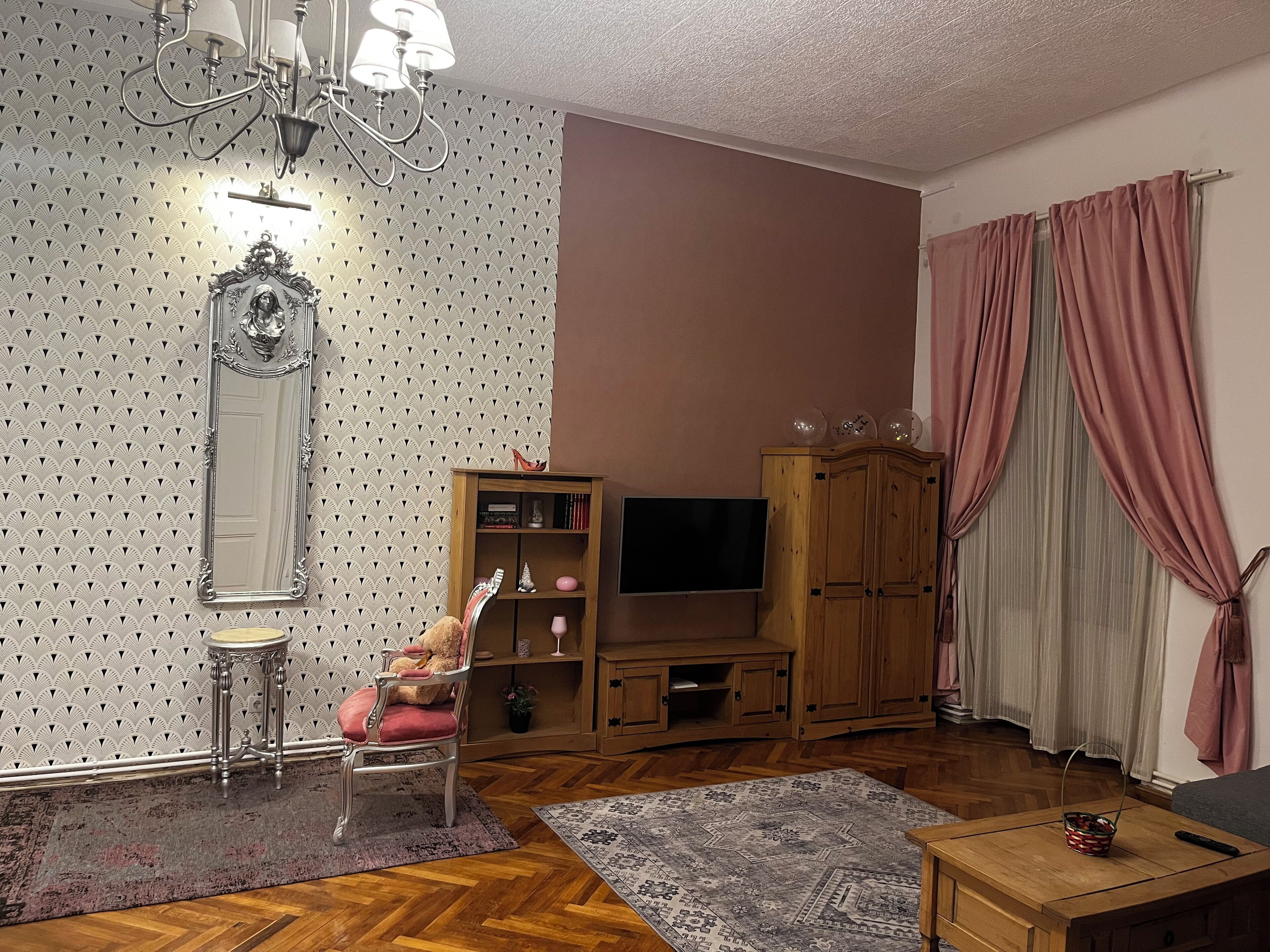 A living room with a patterned wallpaper, a large mirror, a wooden cabinet, a television, and a red chair.