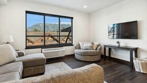 A modern living room with a large window, a flat-screen TV, a sofa, a round ottoman, and a small table.