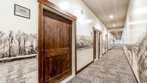 A hallway with a wooden door labeled "GYM," carpeted flooring, and wall art depicting trees and a landscape.