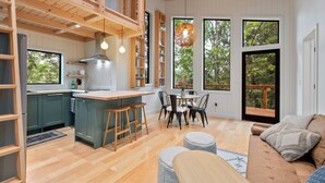 A modern kitchen with wooden beams, a dining area with a round table, and large windows offering views of greenery. - Tree House- Two bedroom plus loft- sustainable living! (Bellingham)