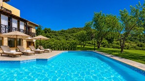 Une piscine entourée de chaises longues et de parasols, elle-même bordée d’une végétation luxuriante et d’un bâtiment aux murs de pierre.