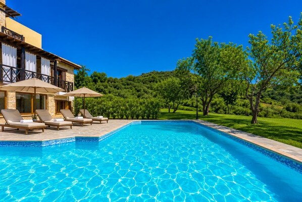 Une piscine entourée de chaises longues et de parasols, elle-même bordée d’une végétation luxuriante et d’un bâtiment aux murs de pierre.