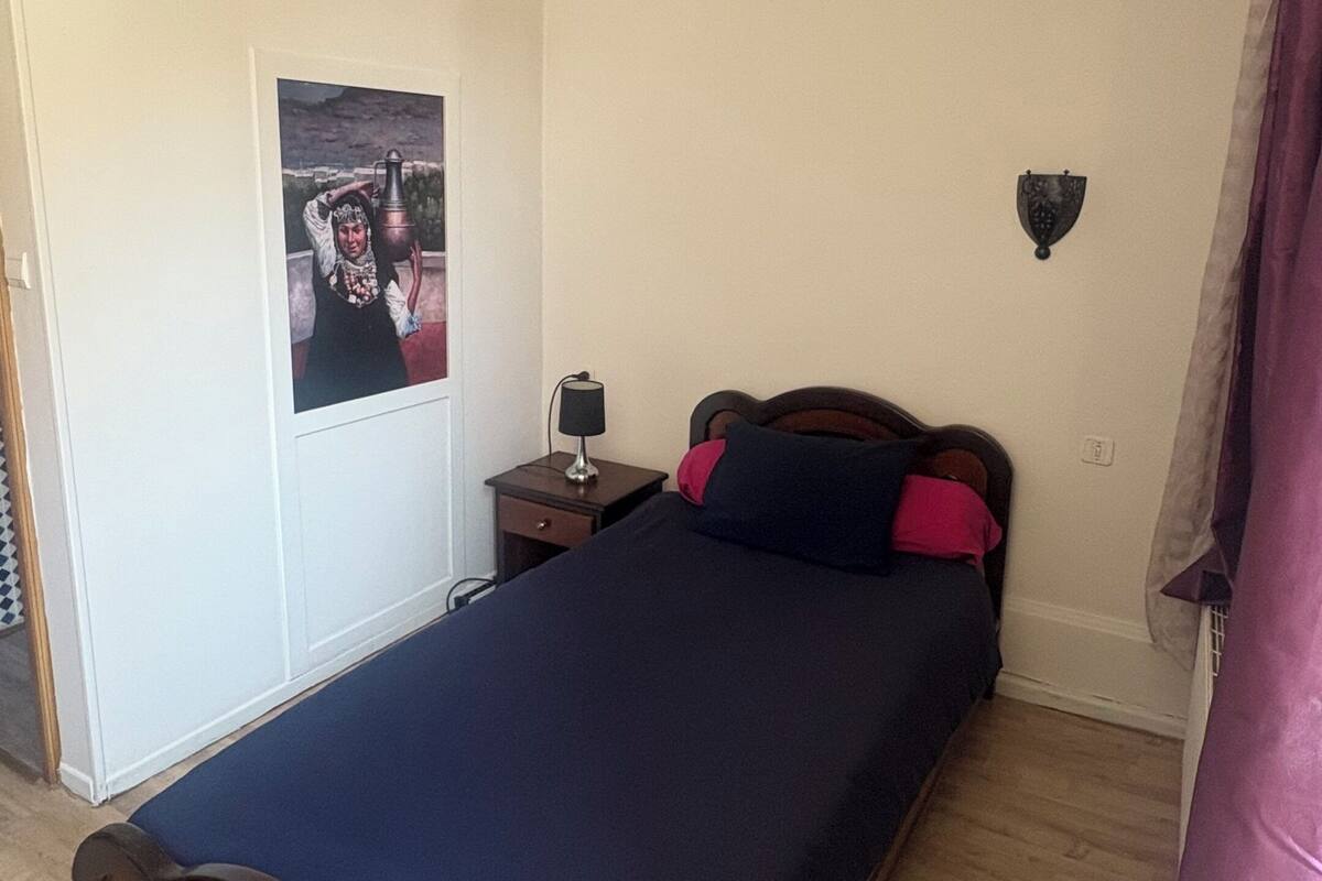 Quarto com cama de madeira, mesa de cabeceira com abajur, luminária na parede e um pôster.