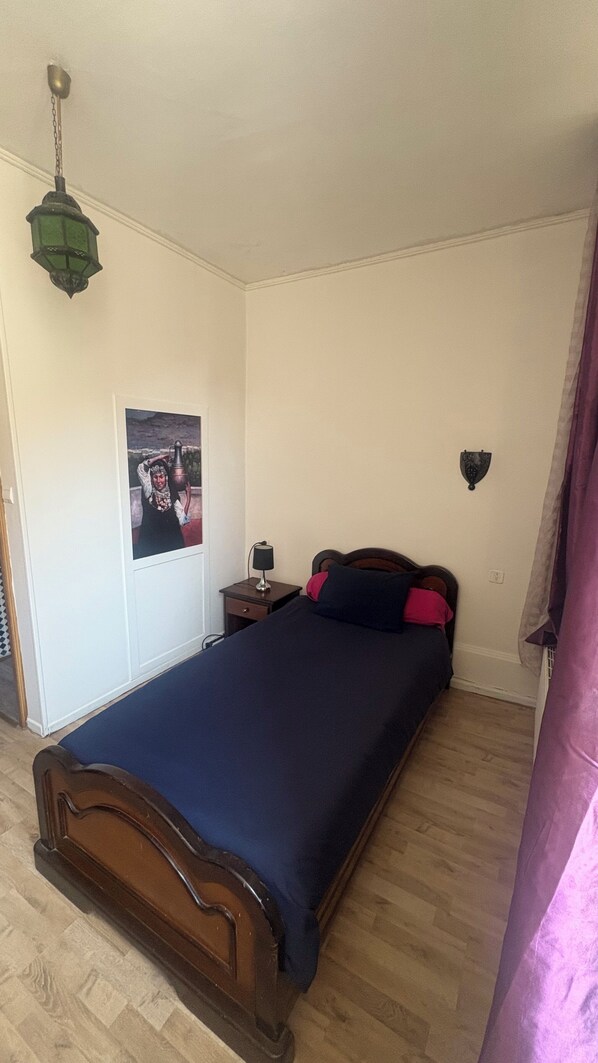 A bedroom with a wooden bed, a nightstand with a lamp, a wall-mounted light fixture, and a poster on the wall.