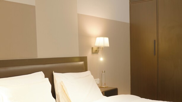 A neatly made bed with white linens, a bedside lamp, and a wooden wardrobe in a hotel room. - Firebrick Garnet House (Gurugram)