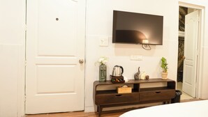 A bedroom with a white door, a flat-screen TV mounted on the wall, a wooden bench, and a bedside table with a vase and a kettle. - Firebrick Garnet House (Gurugram)