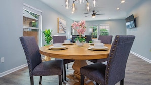 A dining room with a round wooden table, six upholstered chairs, a chandelier, and a ceiling fan.