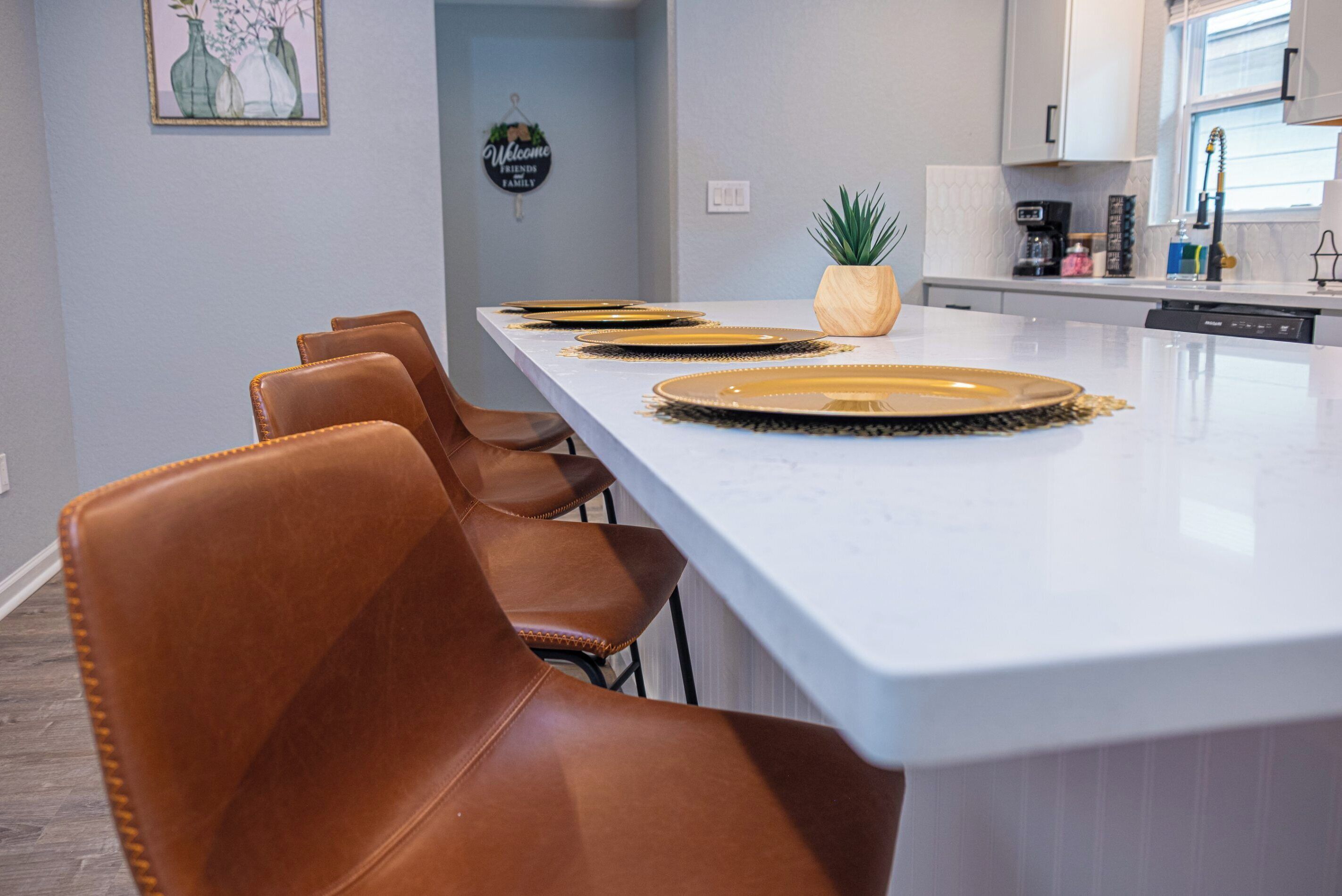 A modern kitchen with a long island, brown leather chairs, and a welcome sign on the wall.