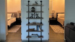 Un interior moderno con una estantería central que exhibe libros y objetos decorativos, flanqueada por dos dormitorios con camas y mesitas de noche.
