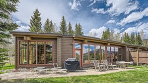 Ein modernes Haus mit großen Glasfenstern, einer Terrasse mit Gartenmöbeln und Blick auf Berge.