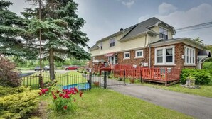 Une maison à deux étages avec un porche à l’avant, une cour clôturée et une voiture rouge garée devant.