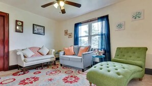 Un salon avec un canapé blanc, un canapé gris et un fauteuil vert. On y trouve un ventilateur de plafond, une fenêtre avec des rideaux bleus et un tapis à motifs floraux.
