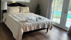 A bedroom with a bed, a nightstand, a lamp, a window with blinds, and a view of a green outdoor area through a glass door. - Stylish house with pool close to strip (Las Vegas)