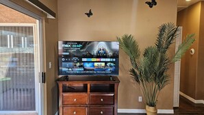 Sala de estar com televisão de tela plana em suporte de madeira, planta em vaso e porta de vidro deslizante.