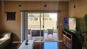 Uma sala de estar com porta de vidro deslizante que dá para um pátio externo, uma televisão sobre um armário de madeira e um vaso de planta.