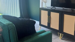 Una habitación con un sofá verde, un televisor negro sobre un mueble negro, y vista a árboles desde afuera.