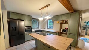 A modern kitchen with green cabinets, a central island, a built-in oven, and a window overlooking greenery. - Mas de Peyre (Le Beaucet)