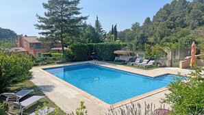 A rectangular swimming pool with a clear blue water surface, surrounded by a stone deck. There are lounge chairs and a small covered seating area with an umbrella. The pool is set in a landscaped garden with various shrubs and trees, and a building with a tiled roof is visible in the background. - Mas de Peyre (Le Beaucet)