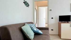 A brown sofa with a teal and blue patterned pillow, a television on a wooden cabinet, and a wall-mounted decorative piece. - Appartamento sul Lungomare by Tempovacanza (Marina di Andora)