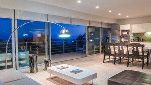 Una sala de estar moderna con sofá, una mesa de centro y una zona de comedor con sillas. Se ve un ventanal con vistas a la ciudad de noche.