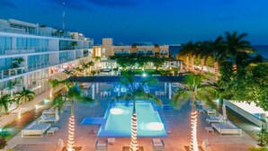 Un área de piscina en un resort, con numerosas tumbonas y una piscina grande, rodeada de palmeras y un edificio con balcones.