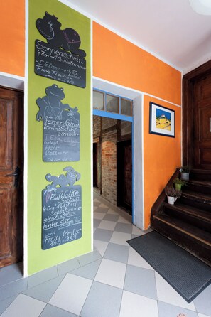 A hotel entrance with a green wall, a chalkboard with room information, a wooden door, and a staircase with potted plants.