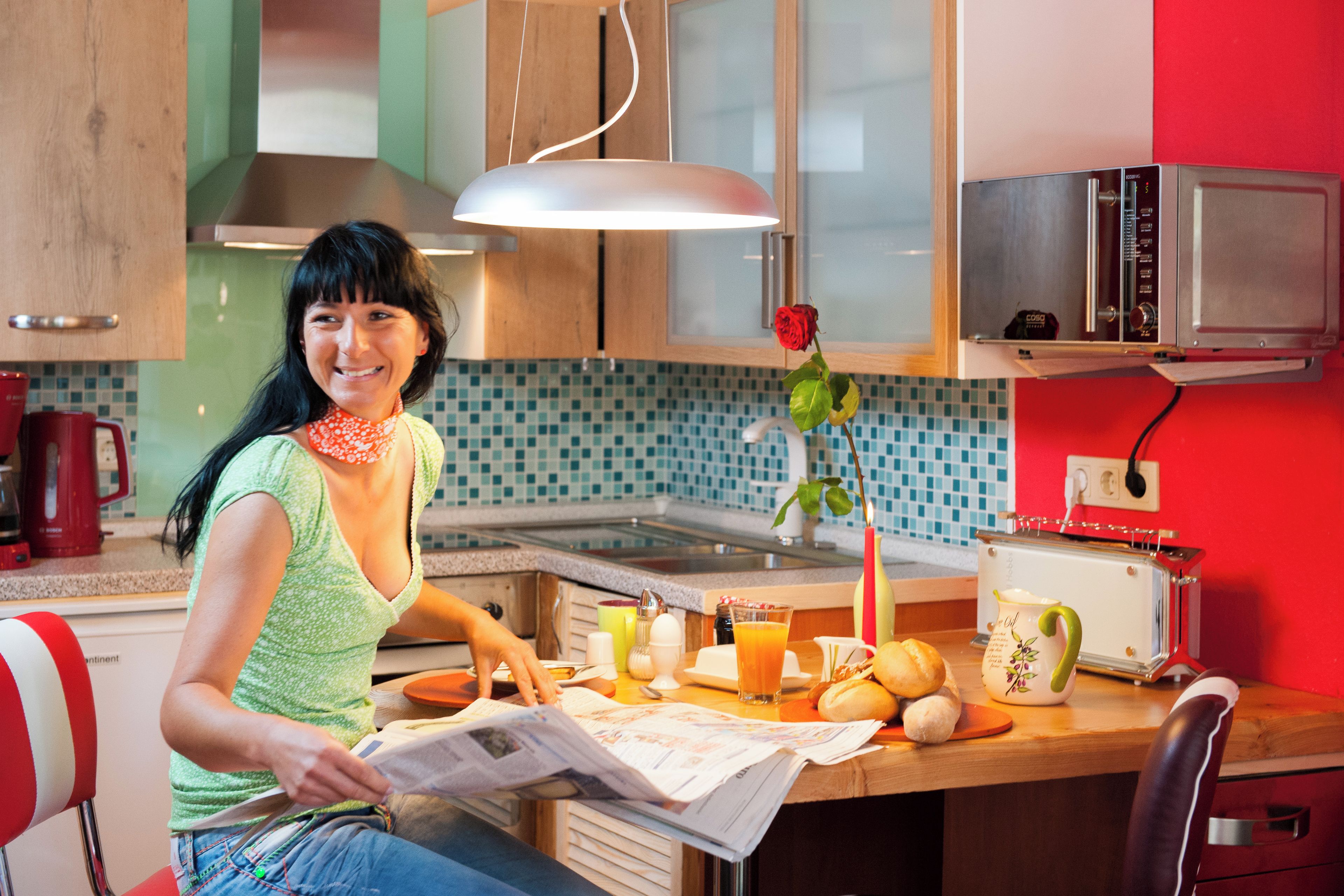 Een keuken met een vrouw die een krant leest, een magnetron, een broodrooster en een roos op het aanrecht.