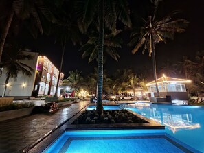 Uma área de piscina à noite, com palmeiras, um edifício com janelas iluminadas e uma piscina azul cristalina.