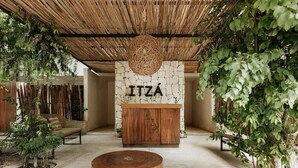 Une pièce avec un mur en pierre, un meuble en bois sur lequel on peut lire le mot « ITZÁ », une suspension lumineuse en fibres tressées et une table ronde en bois.