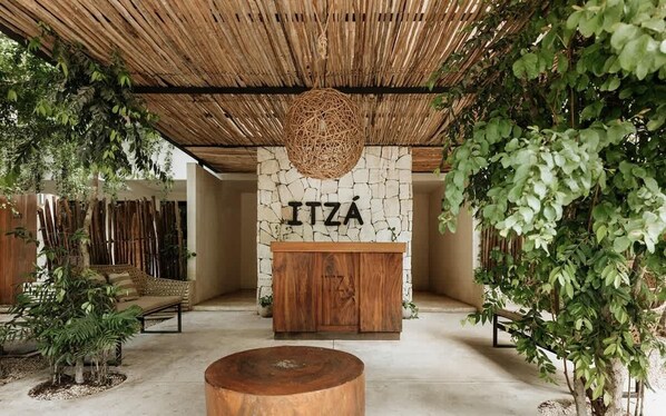 A room with a stone wall, a wooden cabinet with 'ITZÁ' on it, a woven hanging light, and a round wooden table.