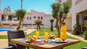 Uma mesa de jantar com pratos de comida e bebidas, uma piscina e um edifício com fachada branca e varandas.