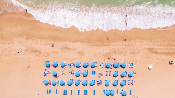 Vista aérea de uma praia com fileiras de guarda-sóis azuis e cadeiras de praia, areal e ondas se aproximando.
